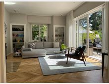 Helles Wohnzimmer mit großen bodentiefen Fenstern, die auf eine Terrasse mit Garten führen. Der Raum ist mit einem grauen Sofa, einem Sessel, einem runden Glastisch und einem hellen Teppich ausgestattet; im Hintergrund befinden sich Regale mit Dekorationsgegenständen.