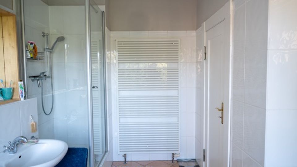 Helles Badezimmer mit weiß gefliesten Wänden, Waschbecken links und einer verglasten Duschkabine. An der Stirnwand steht ein großer weißer Handtuchheizkörper, rechts befindet sich eine weiße Tür; der Boden ist gefliest und mit einem kleinen Badvorleger versehen.