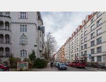 Straßenszene in einem Wohnviertel mit mehrstöckigen Altbauwohnungen auf der linken Seite, die durch dekorative Balkone und Erker gekennzeichnet sind, und modernen Wohngebäuden mit roten Dächern auf der rechten Seite. Geparkte Autos säumen die Straße, während ein Fahrradfahrer auf dem Gehweg unterwegs ist; im Hintergrund stehen kahle Bäume unter einem wolkigen Himmel.