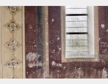 Detailaufnahme einer verwitterten Wand mit verblasster Malerei und eingeritzten Graffiti. Rechts im Bild ist ein kleines Fenster mit horizontalen Glasscheiben sichtbar, umrahmt von abgeblättertem Putz.