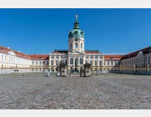 Das Bild zeigt das Schloss Charlottenburg in Berlin, ein barockes Gebäude mit einer zentralen Kuppel, die von einer vergoldeten Statue bekrönt ist. Der Innenhof ist mit Kopfsteinpflaster ausgelegt und durch ein schmiedeeisernes Tor mit verzierten Säulen eingerahmt.