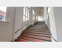 Breite Treppe in einem historischen Gebäude mit weißen Wänden und großen Fenstern, durch die Tageslicht einfällt. Die Treppe hat rote Stufen, schwarze Handläufe und eine Hängelampe über dem oberen Bereich.