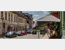 Eine gepflasterte Straße in einer Altstadt mit zweistöckigen Gebäuden im historischen Stil. Links befindet sich ein Restaurant mit der Aufschrift „Gasthaus Schwarzer Adler“, rechts ein Verkaufsstand mit Plüschtieren unter einem Sonnenschirm; geparkte Autos säumen die Straße.