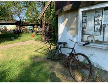 Eine rustikale Außenansicht eines Hofes mit Grasfläche, alten Fahrrädern, und einer weißen Hauswand mit einem Schaukasten, der historische Werkzeuge und Fotos zeigt. Im Hintergrund sind kleine Holzhütten und Bäume sichtbar.