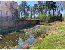 Schmaler, ruhiger Wassergraben mit grünlichem Wasser und Uferbewuchs, daneben eine gemauerte Backsteinmauer. Links steht ein rosafarbenes Gebäude mit hohen Fenstern, im Hintergrund ein Park mit hohen Bäumen und Sträuchern.