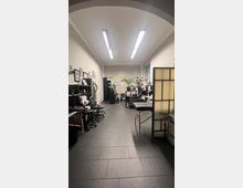 Ein langer Raum mit heller Beleuchtung, der als Tattoo-Studio eingerichtet ist. Der Raum enthält schwarze Arbeitsmöbel, eine Liege, Wanddekorationen mit Bildern und Pflanzen, sowie Trennwände und dunklen Fliesenboden.