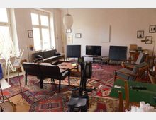 Ein geräumiges Wohnzimmer mit einer Mischung aus modernen und retro-inspirierten Möbeln, darunter ein Sofa, Sessel und Holztische. Der Raum ist mit gemusterten Teppichen ausgelegt, hat helle Wände, hohe Fenster, verschiedene Musikinstrumente und eine Rudermaschine.