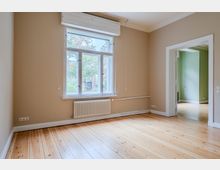 Leerer, heller Wohnraum mit hohen Decken, hellen Holzdielen und beigen Wänden; unter einem großen Fenster mit weißem Rahmen und Rollladenkasten befindet sich ein Heizkörper. Rechts führt eine breite Türöffnung mit weißer Zarge in einen angrenzenden Raum mit grünen Wänden.