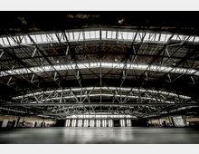 Große Industriehalle mit gewölbtem Dach und sichtbarer Stahlträgerkonstruktion, ergänzt durch breite Fenster oberhalb der Wände, die Tageslicht einlassen. Der Boden ist leer und glatt, die Wände aus Ziegeln und Beton wirken industriell.