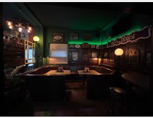 Gemütlicher Pub-Innenraum mit dunkelgrünen Wänden, dunkler Holzvertäfelung und Sitzecken aus Holzbänken um kleine Tische. An den Wänden hängen Schilder und Bilder; warme Kugellampen und ein grüner LED-Lichtstreifen beleuchten den Raum, neben einem Fenster mit Jalousie und einer Tür.