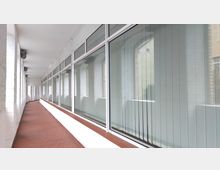 Ein langer, überdachter Flur mit weißen Backsteinwänden und großen Fensterfronten auf der rechten Seite, die mit vertikalen Lamellenjalousien ausgestattet sind. Der Boden ist mit roten Fliesen ausgelegt, und die Decke ist glatt und weiß gestrichen.