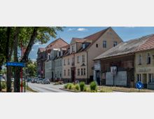 Straßenansicht einer Kleinstadt mit einer Reihe von Gebäuden im historischen Stil, teils renoviert, teils mit abgenutzten Fassaden. Im Vordergrund ein Straßenschild mit der Aufschrift „Straße des Aufbaus“ sowie Bäume und parkende Autos entlang der Straße.