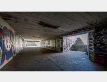 Ein grauer Unterführungsdurchgang mit Wänden und Decken, die von bunten Graffitis bedeckt sind. Im Hintergrund führen zwei Treppenaufgänge ins Freie, die von Tageslicht erhellt werden.