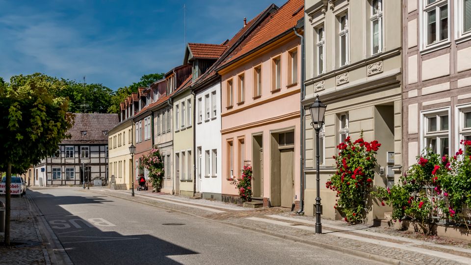 Ruhige Altstadtstraße mit schmaler Fahrbahn, Kopfsteinpflasterrand und einer Reihe historischer Wohnhäuser mit Putz- und Fachwerkfassaden sowie roten Ziegeldächern. Vor mehreren Eingängen stehen Laternen und blühende Rosensträucher, im Hintergrund sind weitere Fachwerkhäuser und Bäume zu sehen.
