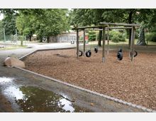 Spielplatz mit Schaukeln aus alten Autoreifen, umgeben von Mulchfläche und Bäumen. Im Hintergrund befinden sich ein Zaun, ein Gebäude mit Graffiti sowie eine Tischtennisplatte.