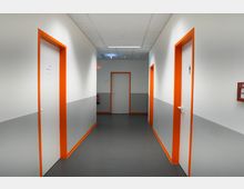 Ein Flur in einem modernen Gebäude mit weißen Wänden, die im unteren Bereich grau abgesetzt sind. Türen mit orangefarbenen Rahmen befinden sich links und rechts, und am Ende des Flurs ist eine weitere Tür zu sehen; eine Feuerlöscheinrichtung hängt an der rechten Wand.