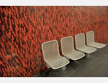 Vier silberne Metallstühle in Gitteroptik stehen in einer Reihe an einer Wand mit rotem Mosaikmuster aus unterschiedlich langen und dunklen Vertikalstreifen. Der Boden ist dunkel und glatt.