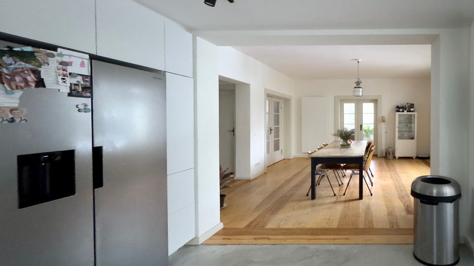 Offener Wohn- und Essbereich mit angrenzender Küche, weißer Einbauzeile und großem Edelstahl-Kühlschrank im Vordergrund. Im Hintergrund stehen ein langer Esstisch auf Holzboden, eine Pendelleuchte sowie eine Glastür mit Tageslicht; der Küchenbereich hat einen grauen Betonboden und schwarze Deckenschienenbeleuchtung.