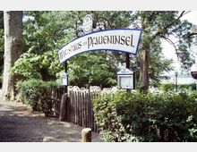 Eingangsbereich eines Biergartens mit dem Schild „Wirtshaus zur Pfaueninsel“ in geschwungener Schrift, umgeben von Bäumen und Sträuchern. Im Vordergrund befindet sich ein Holzzaun, dahinter sind weiße Gartenstühle erkennbar.