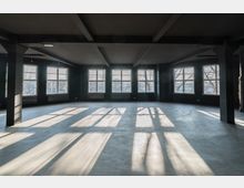 Ein leerer, lichtdurchfluteter Raum mit dunklen Wänden, sichtbaren Deckenbalken und großen rechteckigen Fenstern, durch die Sonnenlicht auf den Betonboden fällt.