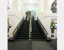 Treppenhaus in einem historischen Gebäude mit breiter, schwarzer Treppe und vergoldetem Handlauf, flankiert von hohen Blumenarrangements. Der Raum ist hell beleuchtet mit weißen Kugellampen und von hohen, weißen Wänden umgeben.