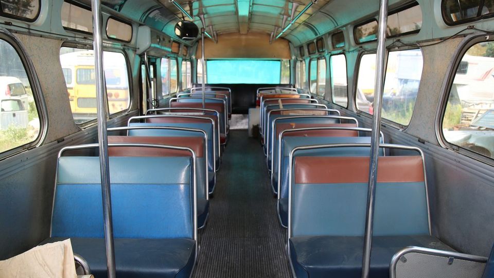 Innenraum eines alten Linienbusses mit zwei Reihen aus gepolsterten Sitzen in Blau und Braun. Die Wände und Decke sind in Metalloptik gehalten, große seitliche Fenster lassen Licht herein.