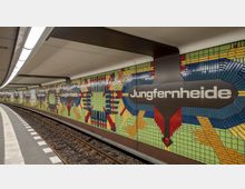 U-Bahn-Station mit dem Namen "Jungfernheide", erkennbar an dekorativen, farbenfrohen Mosaikfliesen an den Wänden in geometrischen Mustern. Der Bahnsteig hat einen gebogenen Verlauf, flankiert von Gleisen und einer hell beleuchteten Decke.