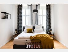 Schlafzimmer mit einem Doppelbett, weißen Bettlaken und Kissen, sowie einer senfgelben Decke und einem passenden Kissen. Im Hintergrund sind große Fenster mit gemusterten Vorhängen zu sehen, flankiert von zwei Nachttischen mit kleinen Lampen, und eine minimalistische Wandregal-Dekoration an der linken Wand.