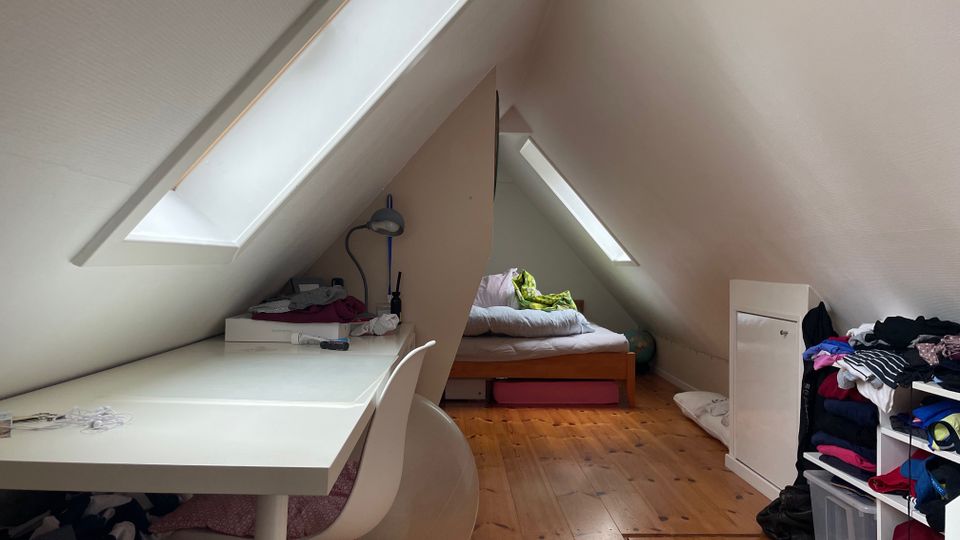 Dachzimmer mit schrägen weißen Wänden und zwei großen Dachfenstern, Holzfußboden und einem kleinen Bett am hinteren Ende. Links steht ein weißer Schreibtisch mit Stuhl, rechts Regale und ein Schrank mit Kleidung und Aufbewahrungsboxen.