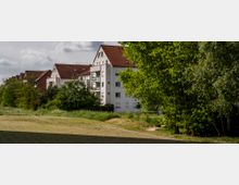 Mehrfamilienhäuser mit weißen Fassaden und roten Ziegeldächern stehen am Rand einer Wiese, umgeben von Bäumen und grüner Vegetation unter einem bewölkten Himmel. Im Vordergrund befindet sich ein gepflasterter Weg und eine Grasfläche.