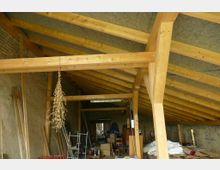 Holzrahmenbau eines Dachstuhls in einem halbfertigen Innenraum mit sichtbaren Holzbalken und einem offenen Dach. Der Raum ist mit Baumaterialien, Möbeln und Werkzeugen gefüllt, während an einer Balkenkonstruktion ein Bündel getrockneter Pflanzen hängt.