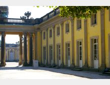 Außenansicht eines gelben barocken Gebäudes mit weißen Fenstern und dekorativen Säulen, das von einem offenen Säulengang flankiert wird; im Vordergrund ein Schild mit der Aufschrift "Shop".