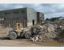 Eine Industriebrache mit beschädigten Wellblech- und Ziegelgebäuden im Hintergrund. Im Vordergrund arbeitet ein gelber Radlader an einem Schutthaufen, umgeben von Erd- und Trümmermassen.