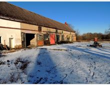 Schneebedeckter Hof mit einem alten, länglichen Gebäude mit Ziegeldach und mehreren Toren und Fenstern. Im Vordergrund sind Fußspuren im Schnee zu sehen, und rechts steht ein kleiner, roter Schornstein auf einem Podest. Im Hintergrund befindet sich eine Wiese mit Bäumen.