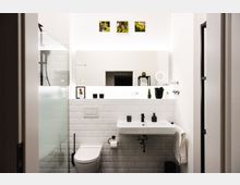 Modernes Badezimmer mit weiß gefliesten Wänden, einem Waschbecken mit Spiegel, einer Toilette und einer Dusche mit teils mattiertem Glas. Dezente Dekorationselemente wie Pflanzen, schwarze Spenderflaschen und kleine Wandbilder mit Moosmotiv sind sichtbar.