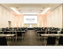 Großer Konferenzraum mit mehreren Reihen von rechteckigen Tischen und schwarzen Stühlen, einem Teppichboden und einer beleuchteten weißen Leinwand an der Frontwand, auf der "Hotel Berlin, Berlin" steht. Auf den Tischen befinden sich Schreibmaterialien und Wasserflaschen.