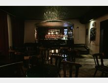 Dimly lit lounge bar interior with wooden floorboards, scattered round tables and chairs, and a leather booth along the wall. At the back, an arched alcove frames a stocked bar counter with hanging lights, a refrigerator display, and a chandelier overhead.