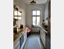 Schmale, zeitgemäß eingerichtete Küche mit weißen Schränken und Holzarbeitsflächen. Im Hintergrund befindet sich ein großes Fenster mit Heizkörper darunter, daneben eine Bank mit rotem Polster.