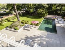 Großer moderner Garten mit rechteckigem Pool, einer Steinterrasse mit Loungemöbeln und einem roten Sonnenschirm. Im Hintergrund grüner Rasen, Bäume und eine Mauer, die das Grundstück umgibt.