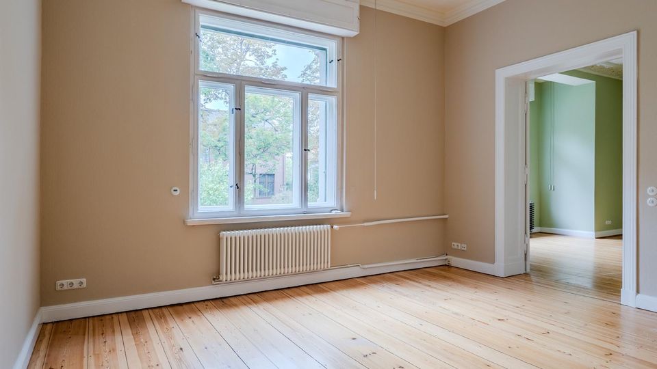 Leerer, heller Wohnraum mit hohen Decken, hellen Holzdielen und beigen Wänden; unter einem großen Fenster mit weißem Rahmen und Rollladenkasten befindet sich ein Heizkörper. Rechts führt eine breite Türöffnung mit weißer Zarge in einen angrenzenden Raum mit grünen Wänden.