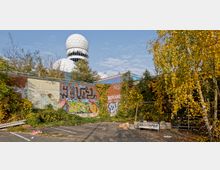 Eine verlassene, graffitibedeckte Backsteinmauer mit dichter Vegetation, darunter gelb gefärbte Herbstbäume. Im Hintergrund ragt eine weiße, kuppelförmige Struktur mit wabenartigem Muster hervor, die zu einem früheren Überwachungsgebäude gehört.