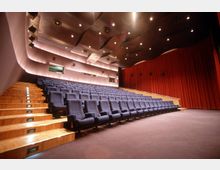 Ein moderner Kinosaal mit ansteigenden Sitzreihen aus blauen Polstersesseln, Holztreppen mit integrierter Beleuchtung und einer Bühne vor einem roten Vorhang. Die Decke ist wellenförmig gestaltet und mit kleinen Deckenspots beleuchtet.