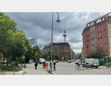 Straßenszene in Berlin mit Blick auf den Fernsehturm im Hintergrund. Links im Vordergrund befinden sich Bäume und ein Grünstreifen, rechts sind mehrstöckige Wohngebäude, während mehrere Personen und Radfahrer die Straße überqueren.