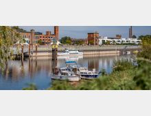 Mehrere Boote liegen in einem kleinen Hafenbecken, das von einer grünen Uferböschung umgeben ist. Im Hintergrund sind renovierte Industriegebäude aus Backstein, moderne Wohnhäuser und ein Schornstein zu sehen, die an ein ehemaliges Industriegebiet erinnern.