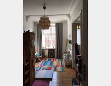 Ein Kinderzimmer mit Holzboden und einer bunten Streifenteppich. Es gibt ein großes Fenster mit grauen Vorhängen, eine hölzerne Hängelampe, Regale mit Spielsachen und ein Globus auf einem Schrank.