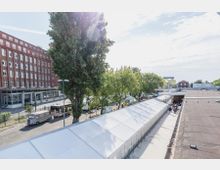 Außenbereich mit einem roten Backsteingebäude links, mehreren Bäumen und weißen Zelten im Vordergrund. Rechts steht ein flaches Gebäude mit dunklem Dach, während im Hintergrund ein Bereich mit weiteren Gebäuden und vereinzelter Vegetation zu sehen ist.