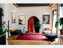 Wohnzimmer mit einer Mischung aus modernem und traditionellem Design: Holzfußboden, ein großer roter Teppich, bunte Sofas und Pflanzen. Im Hintergrund befindet sich ein bogenförmiger Durchgang mit rotem Vorhang, flankiert von abstrakten Wandbildern und hängenden Leuchten.