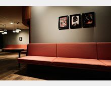 Modernes Lounge-Interieur mit roten Polsterbänken, hölzernem Boden und dimmer Beleuchtung. An der grauen Wand hängen drei abstrakte Kunstwerke in schwarzen Rahmen.