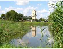 Ein Gewässer mit Schilfbewuchs im Vordergrund, dahinter ein mehrstöckiges Gebäude mit heller Fassade, das auf Stelzen steht. Umgeben von grünen Bäumen und Wiesen mit vereinzelten Anhängern im Hintergrund bei sonnigem Wetter.