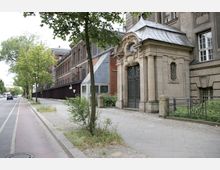 Straßenszene mit historischen Gebäuden, darunter ein renoviertes Backsteingebäude und ein rechteckiges Torhaus mit kunstvoll verzierten schmiedeeisernen Toren und einem dekorativen Giebel. Links verläuft eine Straße mit Fahrradweg, flankiert von Bäumen und Gehwegen.